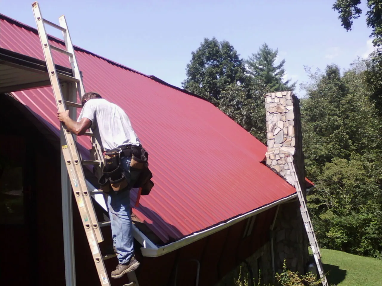 Roofer on a ladder beside a red metal roof and stone chimney — Soffit & Fascia Repair by Roofing Brothers in Grant Park